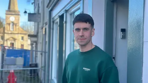 Eóin McAlonan, a man with short, dark hair, looking to the camera on a street in Ballycastle. He is wearing a green crew-neck sweatshirt. There are steel barriers behind him and a church with a clock tower in the background.