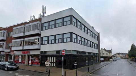 The corner three-storey building. The ground floor is black, the top two area white with windows. There is a Wimpy restaurant to the left of the building. A road runs down the right side.