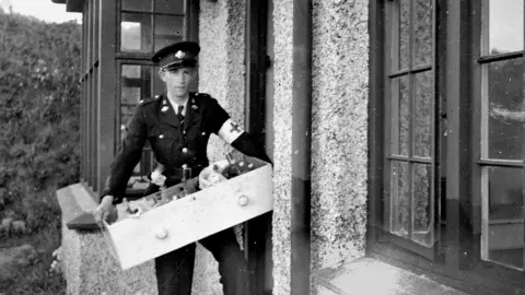 St John Ambulance and Rescue Service A black and white photo of a young man in a military-style uniform with a white armband on with a cross on it. He is at the door of a house, bringing in a drawer of medical equipment.