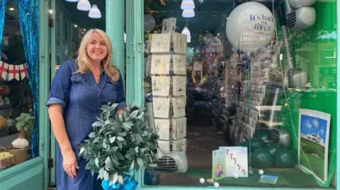 BBC/Angela Ferguson A woman with long blonde hair and wearing a short-sleeved denim dress is smiling while standing outside the door of a shop. She is next to a window display which includes inflatable golf clubs, inflatable golf balls, gold balls, a painting of the Royal Birkdale and a children's book about golf, as well as a larger white balloon with 'It's back', 'The Open' and 'At Royal Birkdale' on it.