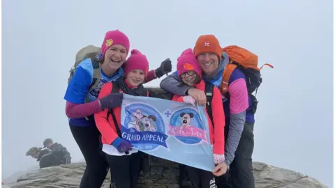 BBC Kara pictured with her mum Sarah, sister Isobel and Dad Stephen at the summit of Snowdon. They are holding a grand appeal flag.