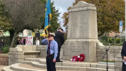 A man lays a poppy wreath at the memorial in St Ouen during a Remembrance Sunday service. Another man who is wearing a blue uniform is holding a blue flag.