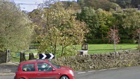 A small red car drives past a small park surrounded by dry stone wall.