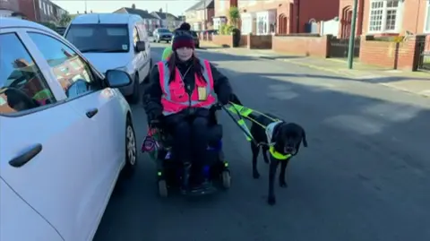 A woman sitting in an electric wheelchair in the middle of a road, with a guide dog beside her. Cars are parked to the left, with houses to either side. She is wearing a purple bobble hat, dark clothes and a pink hi-vis jacket. The dog is black and wears a bright green harness and lead.