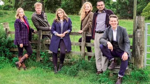 BBC SCOTLAND/BBC STUDIOS Landward presenters Arlene Stuart, Cammy Wilson, Anne McAlpine, Rosie Morton, Shahbaz Majeed, Dougie Vipond, dressed in fashionable country clothes, pose around a fence in a lush green field