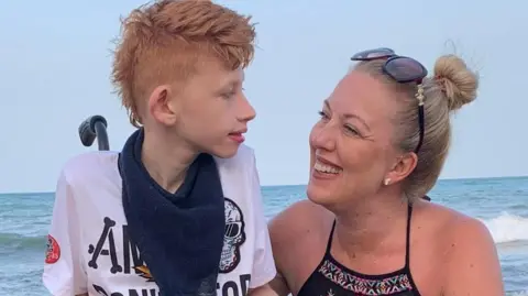 Carl Stockton Mother Claire Stockton has blonde hair tied back and she is wearing a black summer dress. She is sat down smiling at her son who is a wheelchair user and is now 20 years old. Her son is disabled and has a rare condition called ReNU syndrome. He is wearing a white t-shirt with a blue scarf and is looking back at his Mum, Claire. Both are sitting in front of the sea.