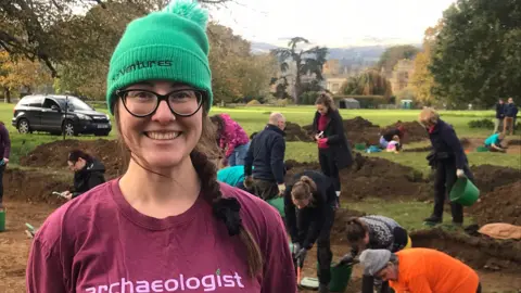 Harriet Tatton stands smiling at the camera as people behind her carry out the dig. Sudeley Castle can be seen in the background. Ms Tatton is wearing a burgundy t-shirt with "archaeologist" written on it in a pink font, and a green beanie with a black logo on it. Her brown hair is in a plait and she is wearing black glasses.