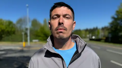 HGV driver Keith Leake looks into the camera. He is wearing a grey jacket and a blue T-shirt. He is standing in front of the junction of Packington Hill and Pritchard Drive.