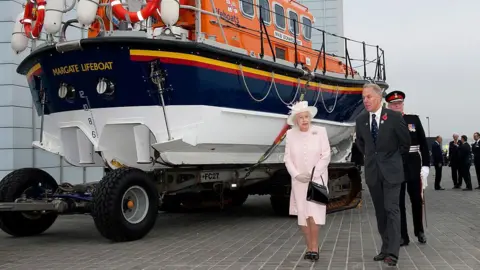 Getty Images Queen Elizabeth II walking past a blue, white and orange RNLI lifeboat which says "MARGATE LIFEBOAT" on the back.