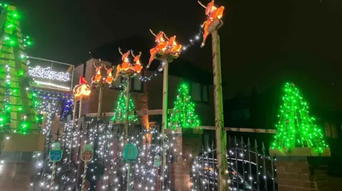 Paul Molyneux Several illuminated reindeer figures are mounted on tall poles, appearing to “fly” above the display. Below them, green light-up trees and white string lights decorate the fence. The scene adds a dynamic touch to the Christmas theme.
