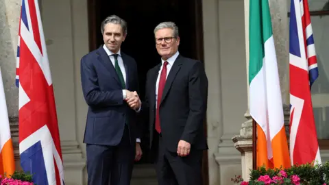 Reuters Taoiseach Simon Harris shaking hands with Prime Minister Sir Keir Starmer outside Farmleigh House in Dublin. There are Irish and Union Jack flags on either side of the two leaders. 