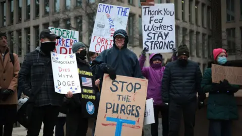 A protest in front of  Office of Personnel Management in February 2025, with people holding signs reading 'stop the purges' and 'Thank you Fed workers stay strong'. 