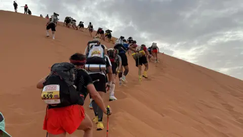 Adam Van de Velde A group of runners in a long line snaking up a sand dune carrying rucksacks on their backs, in the foreground a man is wearing red shorts and a black t-shirt and carrying a stick to help him climb the sand dune
