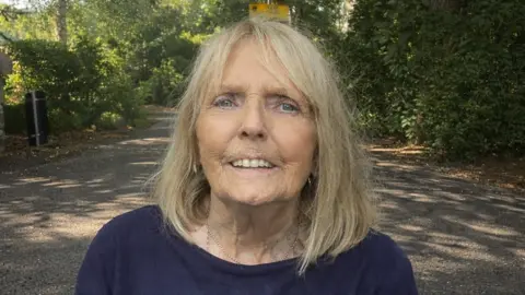 Jackie Whitehurst is looking at the camera and smiling. She has blonde, shoulder-length hair and is wearing a blue sweatshirt. She is in a park on a sunny day. 