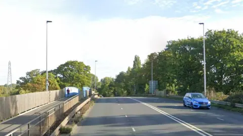 Google image of Bridge Road in Exeter. A blue car is travelling on the carriageway. It is a sunny day with blue skies. Green trees and lampposts line the road. A footpath runs along the left side of the road. 