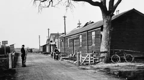 Brian Turpin Single story buildings by the side of a road. A man with a rifle is stood by a fence post and another is riding a bike in the black and white image. There is a large tree in the foreground.