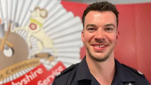 A man with dark hair and a dark beard stood in front of Derbyshire Fire and Rescue insignia.