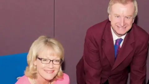 Wendy Austin sitting at a desk smiling up at the camera. She is wearing a pink top. Seamus McKee is standing leaning over with his hands over the desk. He is wearing a suit and tie.