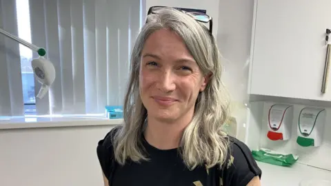 Dr Thomas is in a white, clinical space, with cupboards, a window and a sink behind her. She is wearing a black t-shirt with gold lightning bolts, and is smiling. She has shoulder-length grey hair. It is a head and shoulders shot.