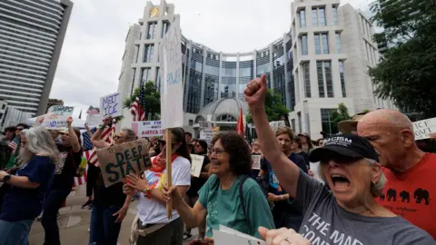 Getty Images A group carrying signs with slogans such as "Melt Ice" and "This is the Deep State" march outside a large building