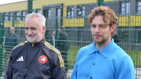 Central Bedfordshire Tom Grennan pictured at the school new 3G sports pitch. He wears a bright blue long sleeve shirt with black buttons. He has curly hair, a gold ear piercing and a tattoo on his neck. He is stood to the right of a grey haired man wearing a navy blue tracksuit top with a round UEFA badge on. 