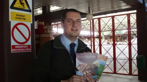 Supplied Lloyd Evans in a shirt and tie. He is in a building in Croatia, holding Jehovah's Witness pamphlets.