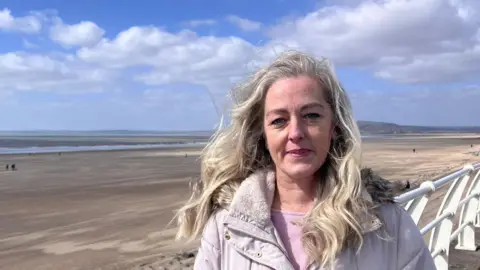 Jo Phillips smiles at the camera. She is standing on a promenade in front of a beach