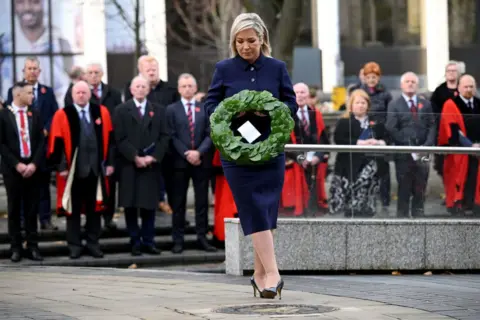 Getty Images Michelle O'Neill in navy skirt suit and black heels, laying a green laurel wreath at remembrance event in Belfast City Hall, out of focus crowd in background watch on in dark clothes