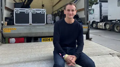 Jim Glennie is wearing a blue long sleeved top and jeans and is sitting on a platform on the back of a lorry.