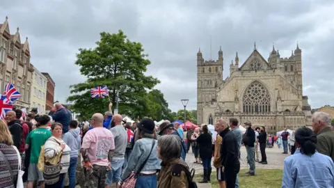 A crowd of people stood in front of Exeter Cathedral green. Some people are waving Union Jack flags.