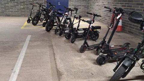 A row of scooters, in a room, with grey brick walls, and three traffic cones to the left. They are all black and standing up 