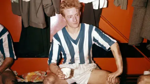 Getty Images Denis Law drinks a cup of team after a Huddersfield Town game in 1957 when he was 16. He wears a blue and white striped football strip with white shorts. He appears to be sitting in the changing room, with clothes hanging on pegs behind him. 