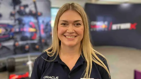 Sam Read/BBC A smiling blonde woman at an event, with a disassembled F1 car behind her.