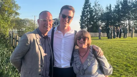 PA Media Three people stand smiling in a sunny field. Arthur's father is seen in a grey blazer on the left, Arthur is pictured in a white shirt and braces in the centre, and his mother is seen in a patterned dress on the right.