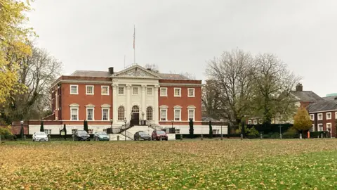 Warrington town hall stands in front of a large grassy area covered by yellow autumn leaves. The red-brick building has four white columns dominating its centre