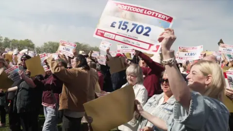 A blonde haired woman in the foreground holds a mock cheque up above her head for £15,702. In the background crowds of people hold up their own amounts - such as £7,851 and £23,553. 