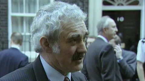 John Burton is speaking to someone outside of the 10 Downing Street. He has short, white hair and a black moustache. He is wearing a blue pin-stripe suite and a shirt. There are people in suits standing behind him too.