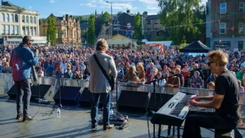 BwD Council Generic image from the back of the main stage showing a male singer, a guitarist and keyboard player watched by hundreds of people crowded before the stage in the sunshine