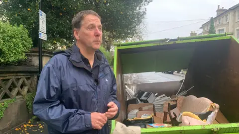 BBC Dan Thomas, wearing a rain jacket, stands next to a skip containing ruined possessions on 8 November 2023