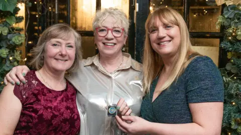 Health Innovation West of England Three women posing for a photo following the award show. The woman on the left has shoulder length brown hair with grey streaks, and is wearing a red lace blouse. In the middle is Ann Remmers, with short white curly hair, wearing a silver button up blouse and red framed glasses. She is smiling and holding the award, which is a small metal coin in a plastic square case. On the right is another woman with long blonde hair, wearing a blue sparkly top . 