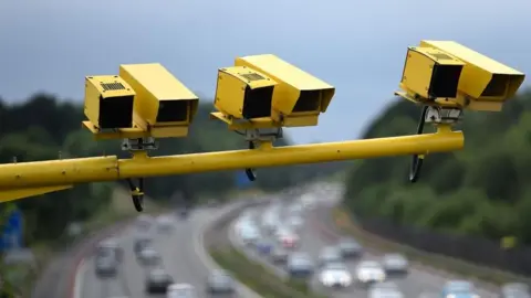 PA Media Several yellow speeding cameras sit on a yellow pole, high above the lanes of the M3 motorway.
