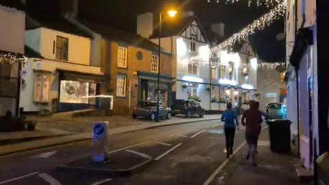 Two women and a man run up the pavement of a town centre street at night. A pub and two shops are on the other side of the street with two cars parked in front of them. The street is lit by a street lamp and Christmas lights strung from one side of the street to the other. One of the shops is also decorated with Christmas lights and a Christmas tree in the window.