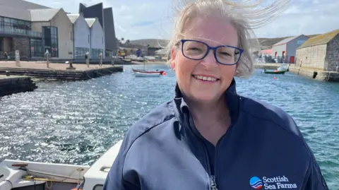 A windswept Anne Anderson photographed from the chest up in a marina setting. She has grey hair - tied back - blue-rimmed glasses and is smiling at the camera. She is wearing a blue jacket with a "Scottish Sea Farms" logo on the left hand side.