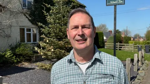Charlie Stubbs/BBC There is a man in a teal chequered shirt, with the top two buttons undone, and a white T-shirt below. To his left is a pub with a white cobbled wall and overgrown trees. The window to the pub has its blinds closed.