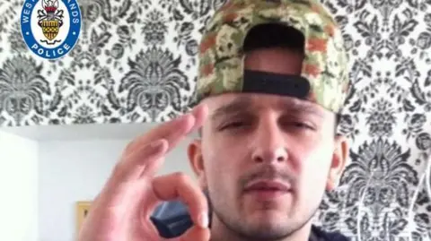 Police handout A man  wearing a khaki baseball cap worn backwards looks into the camera. he has a bit of a moustache and is holding his thumb and forefinger together. He is inside a property which has a black and white wallpaper pattern behind him.