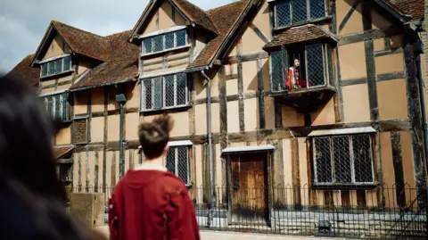 A performance takes place at Shakespeare's Birthplace, the playwright's childhood home. An onlooker stands in front of the half-timbered house and there is an actor at an open window above.