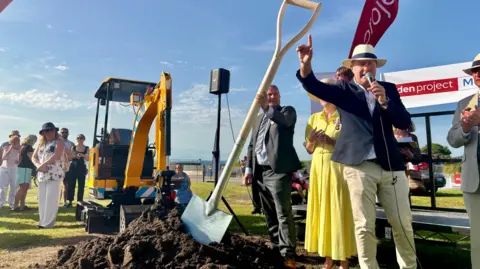 Robbie MacDonald/LDRS The Eden Project Morecambe groundbreaking ceremony with Eden chief executive Andy Jasper, hand raised talking into a microphone, Amanda Parker, the Lord Lieutenant of Lancashire, wearing a yellow outfit and John Pye wearing a grey suit holding a giant seven foot-long spade made by Lancaster & Morecambe College students. Onlookers watch the spade being put into the mound of soil. It is a sunny day.