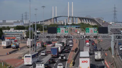 National Highways Cars and lorries queueing on the approach to the Dartford Crossing. The QE2 Bridge is visible and several pylons in the background. There are blue skies and clouds.