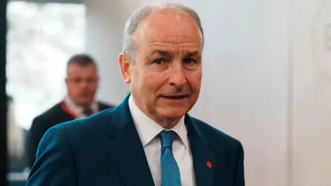 Reuters Micheal Martin wearing a blue suit, white shirt and blue tie. He has grey hair and is looking at the camera.