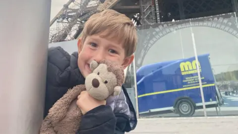 Family handout A young boy peeking round the corner of a grey post smiling in a cheeky way and holding a brown monkey teddy up to his cheek. He has dark blond hair and a fringe and is wearing a black coat. The bottom of the Eiffel Tower is in the background, as is a reflection of a blue van.